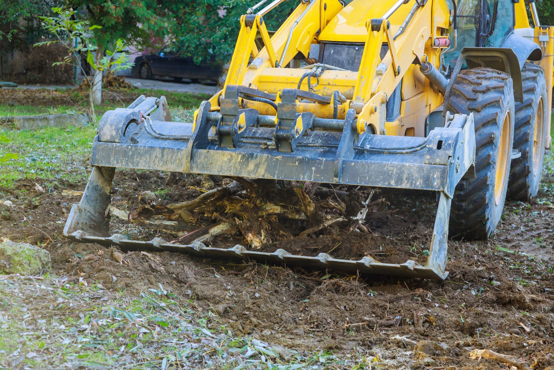 A yellow backhoe tractor sits on a grassy field, using its front loader to dig into the ground and pull up tree roots.