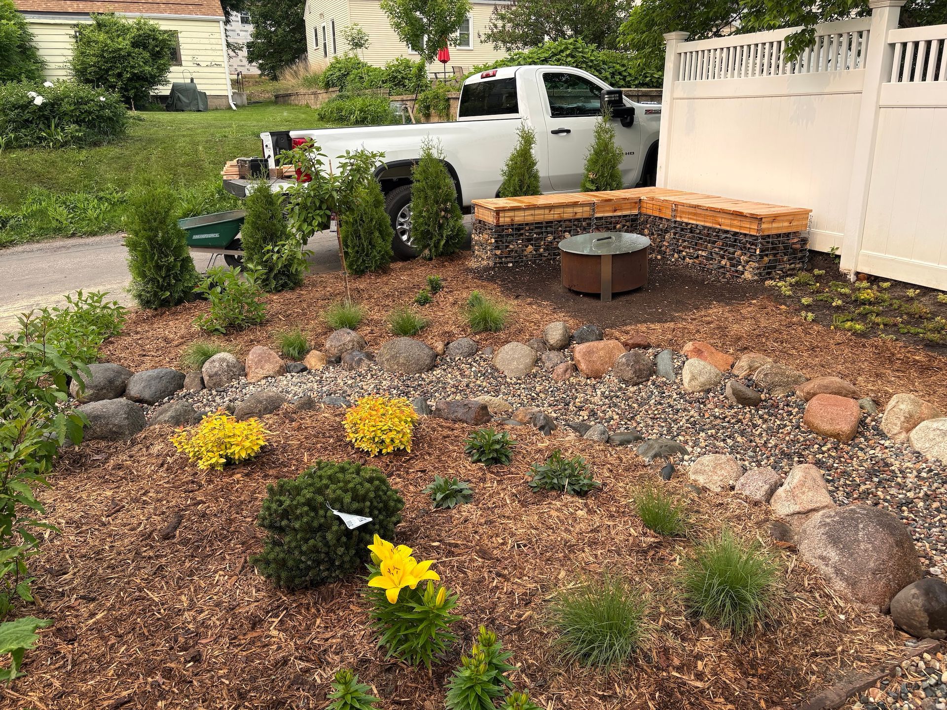 A yard with mulch beds, a dry creek bed of river rocks, small shrubs, yellow flowers, and a stone bench around a fire pit.