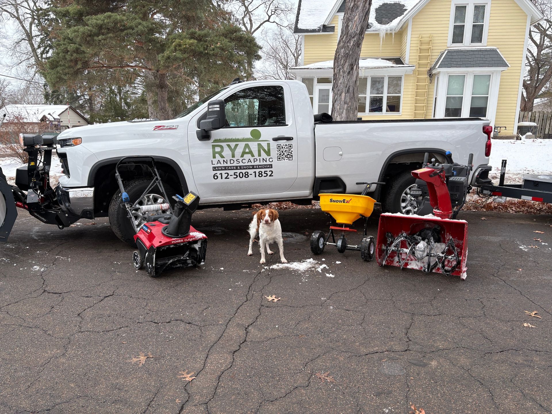 A white pickup truck with a snowplow parked in a driveway next to a dog, two snowblowers, and a yellow salt spreader.