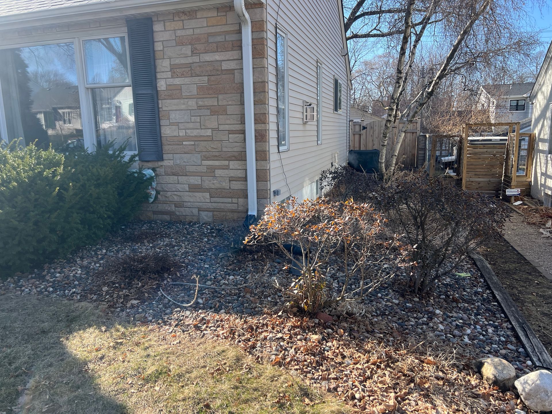 A tan house with stone veneer siding features a side yard with landscaping, dry bushes, and a gravel ground cover.