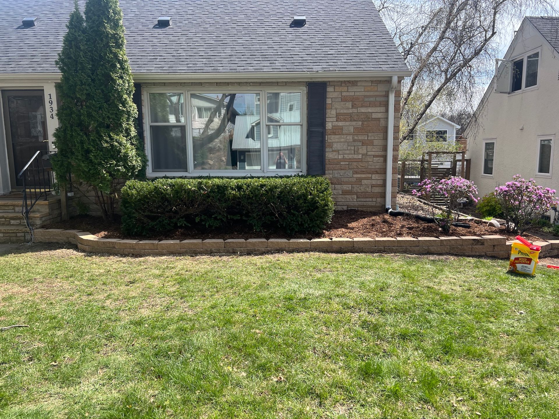 A stone-faced house with a front garden bed featuring a manicured hedge, mulch, and small flowering bushes.