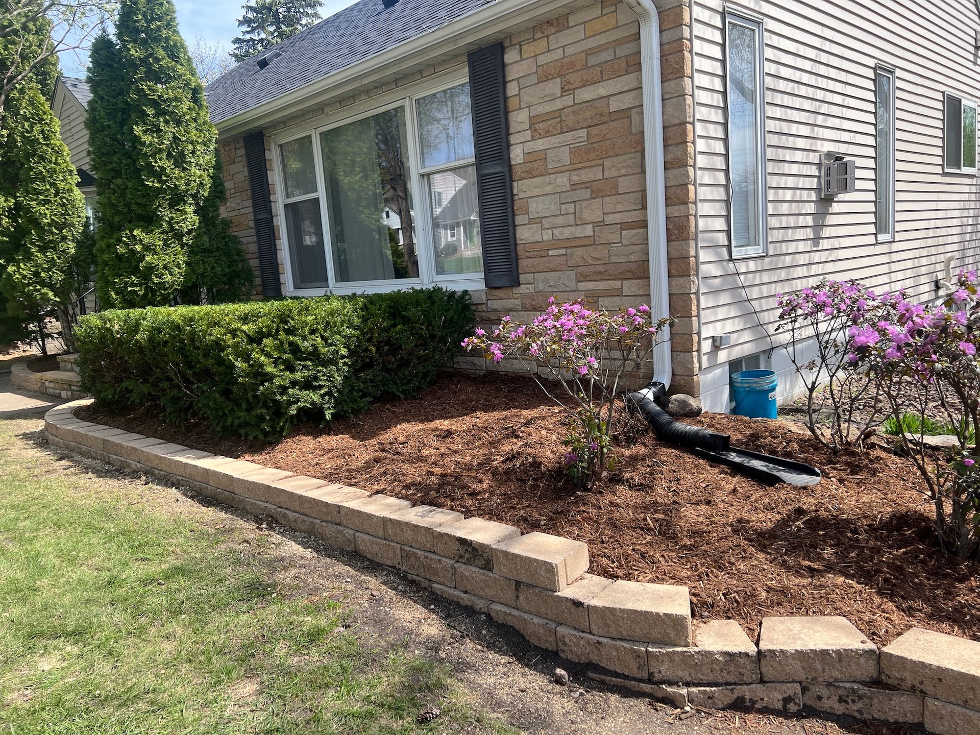 A stone retaining wall surrounds a mulched garden bed with shrubs and purple flowers in front of a tan-sided house.