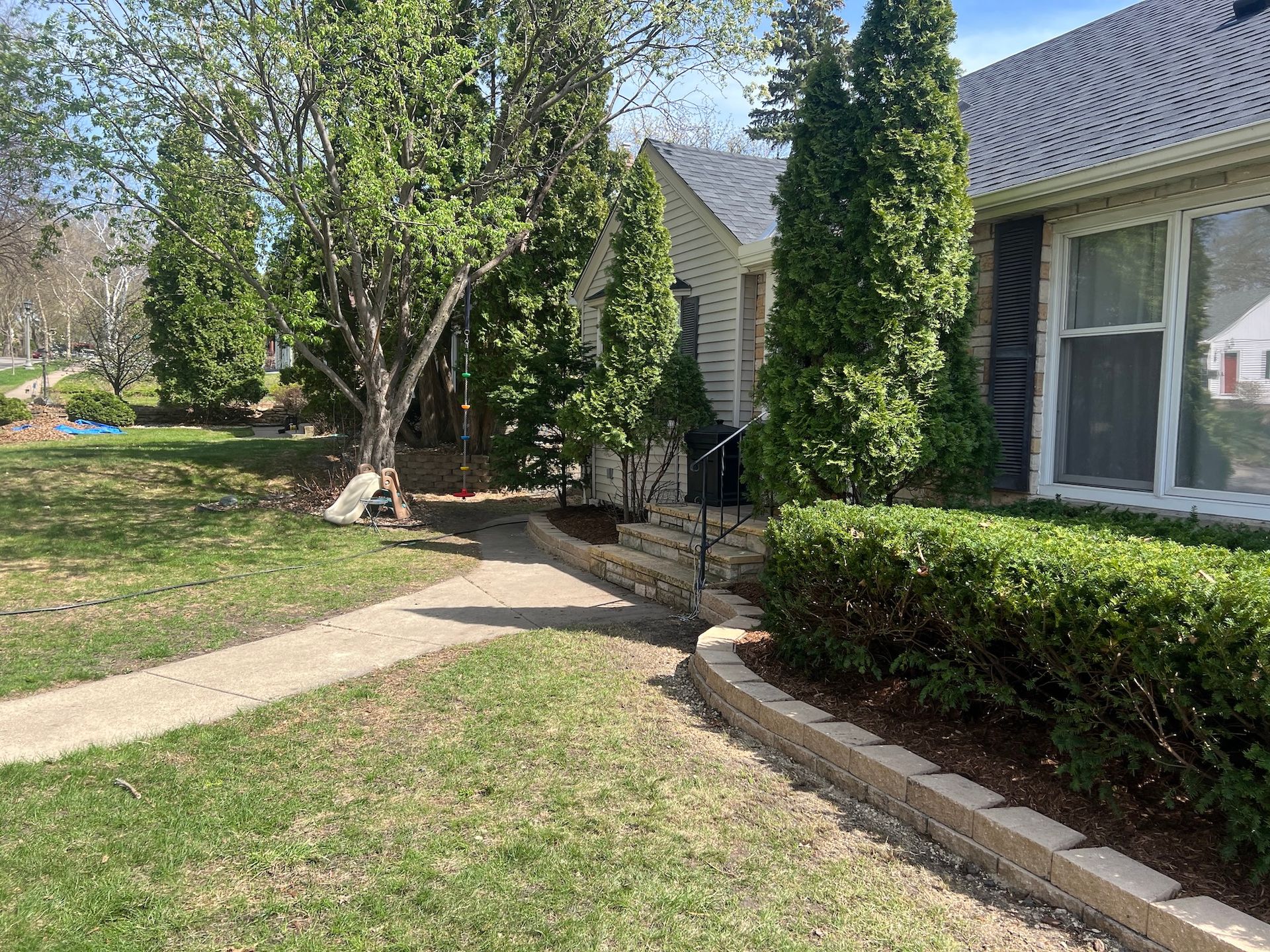 A beige house with dark shutters, a concrete path, a stone retaining wall, and tall evergreen trees in a front yard.