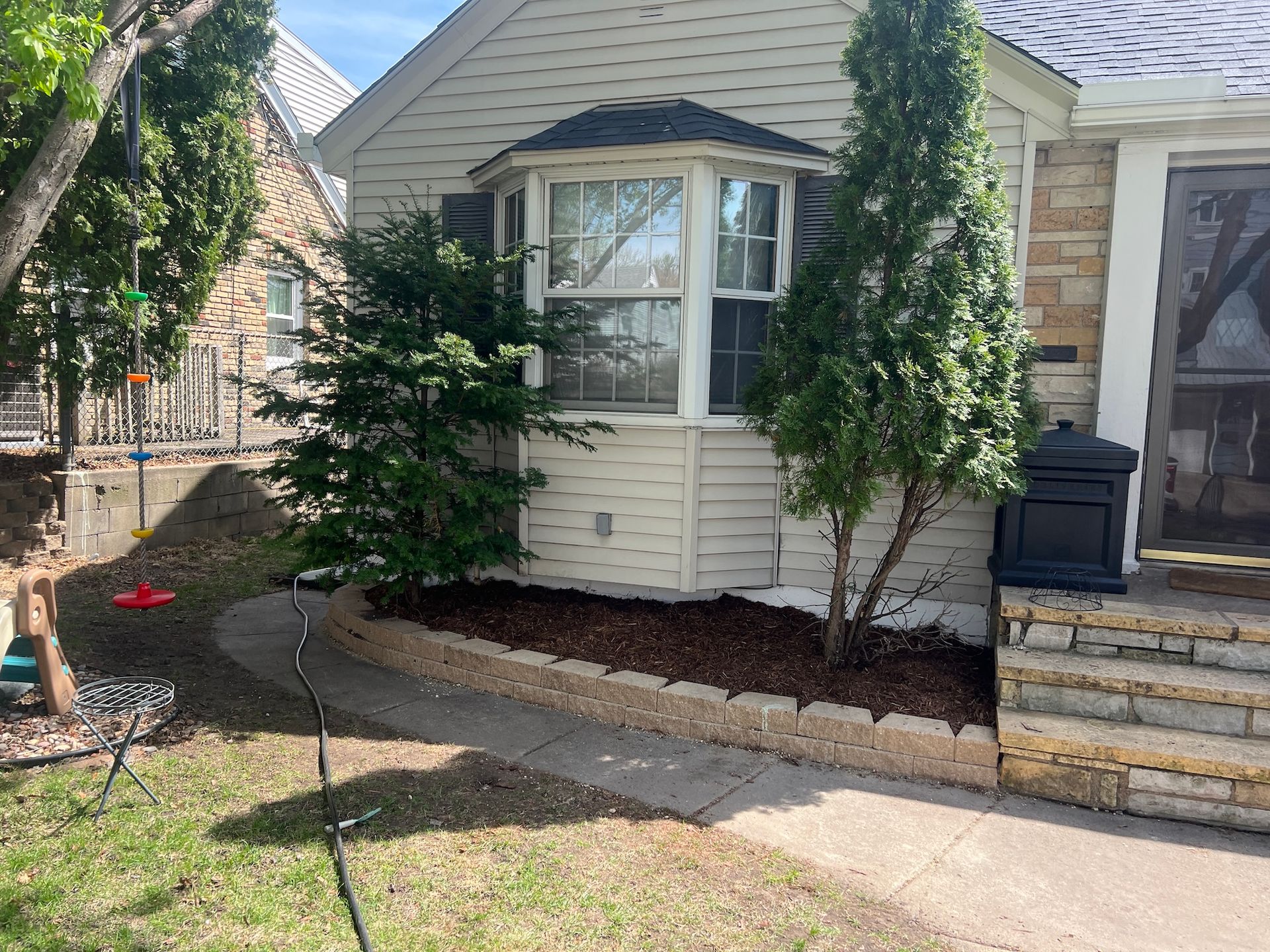 A home exterior features a bay window, a small garden bed with mulch and two evergreen trees, and stone steps.
