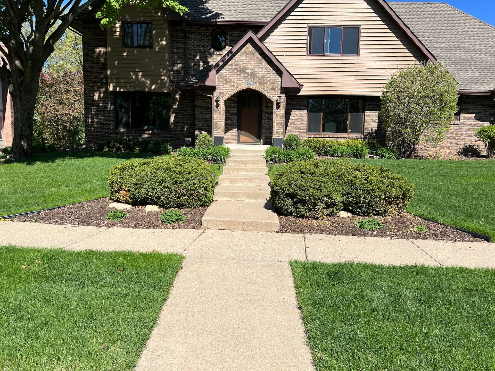 A two-story brick and wood-sided house with a stone entryway, green lawn, and a sidewalk leading to the front door.