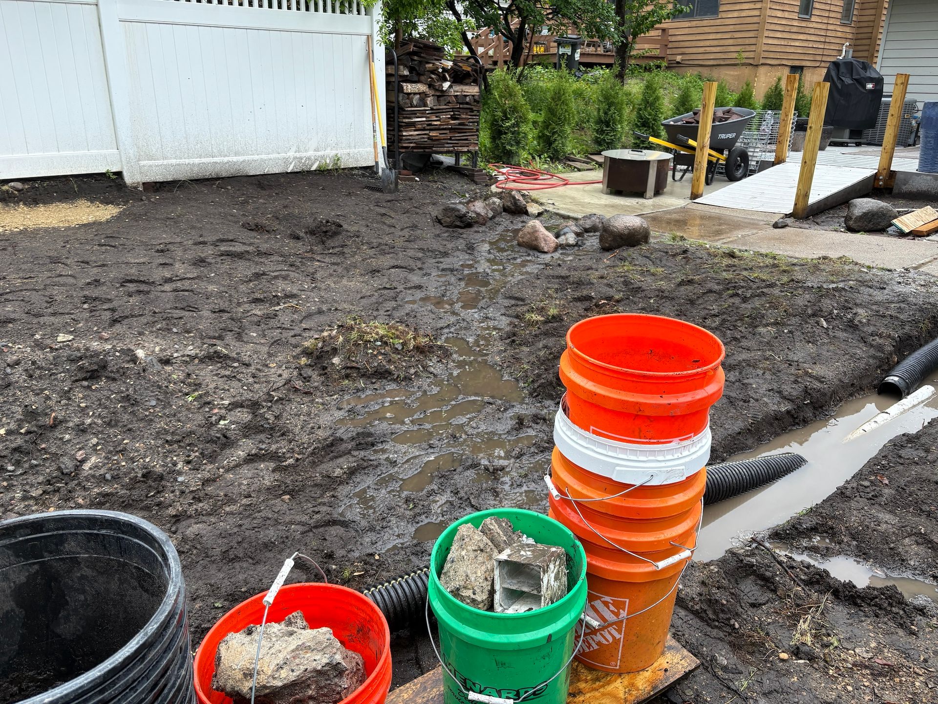 A muddy backyard construction site with a stack of buckets, rocks, and an open drainage trench.