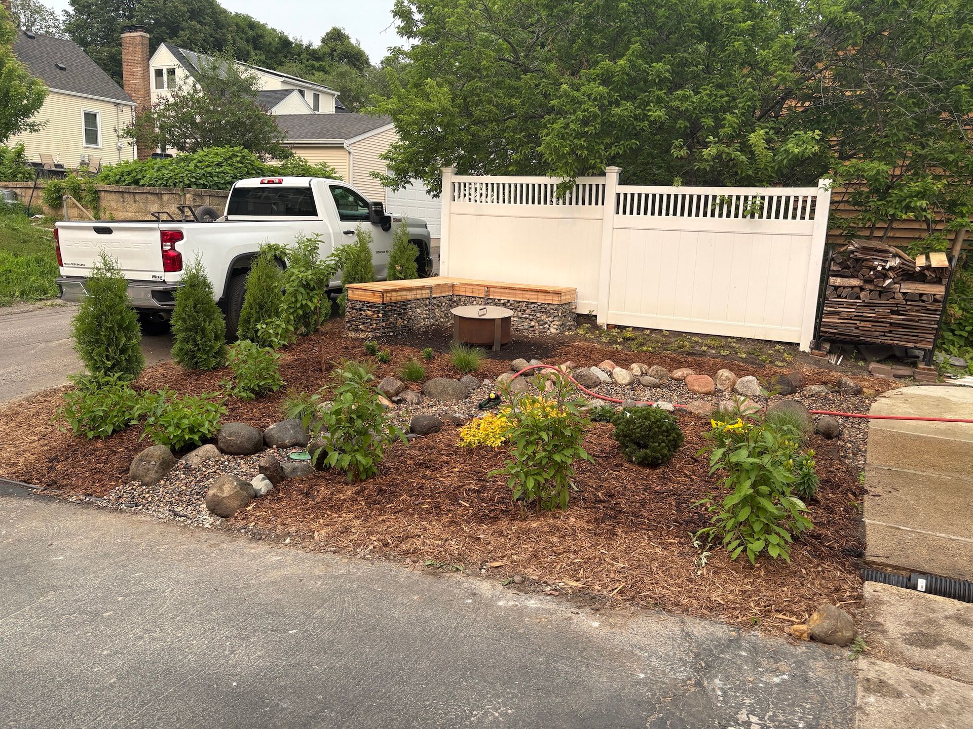 A landscaping project with mulch, stones, and young shrubs in front of a white privacy fence, with a truck parked.
