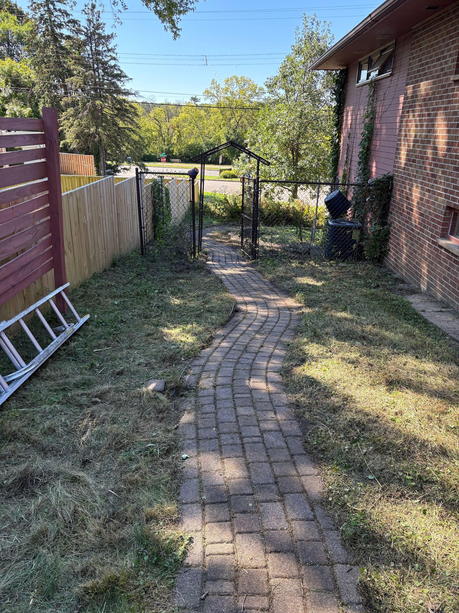 A brick pathway leads between a red wooden fence and a brick house toward a wire gate in a sunny, grassy backyard.