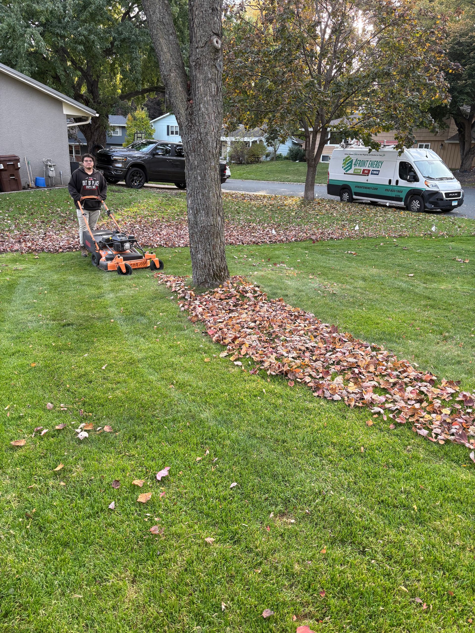A person pushing an lawn mower on a grassy lawn covered in scattered autumn leaves near a house and a service van.