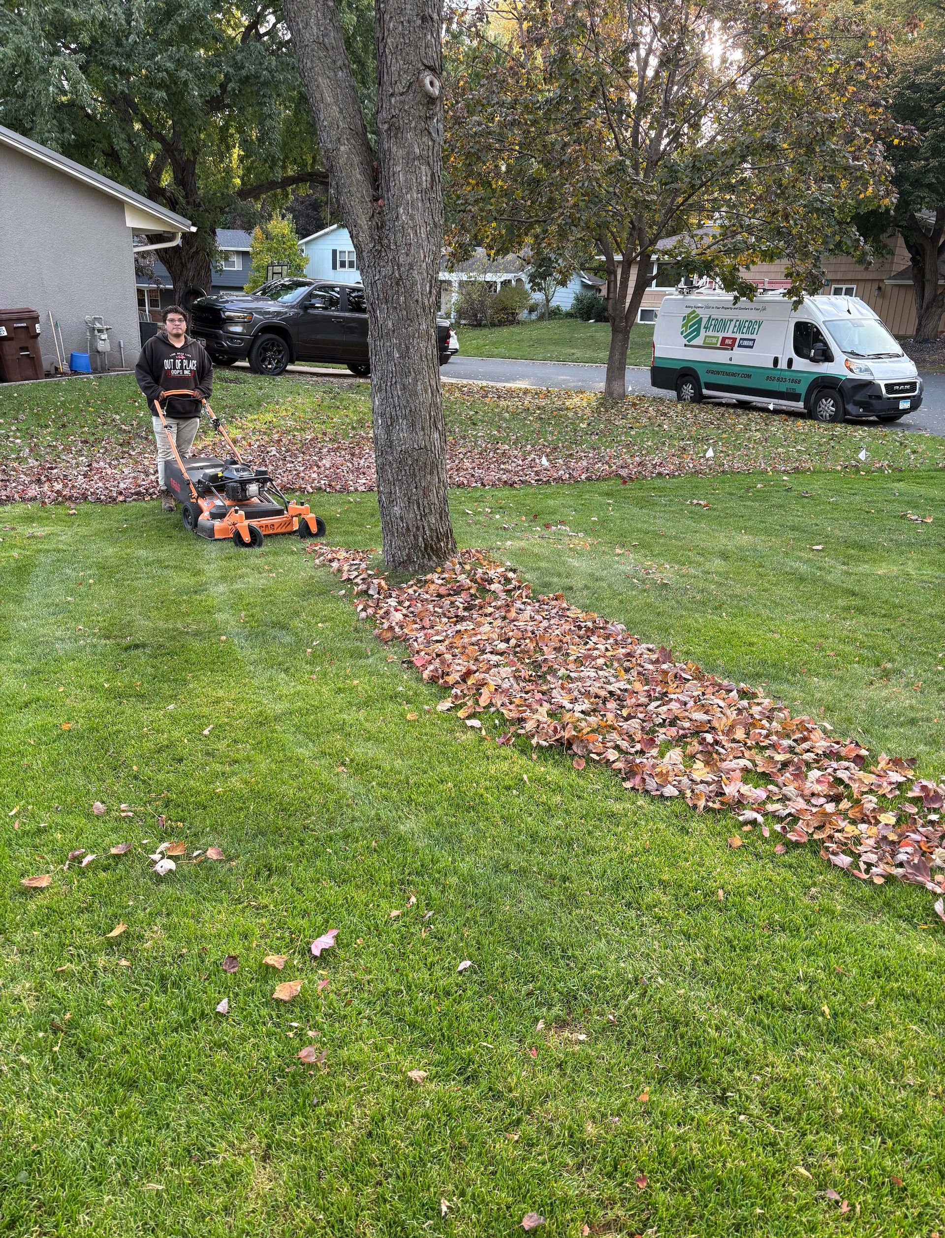 A person mows a lawn covered in fallen leaves in a residential yard near a parked work van.