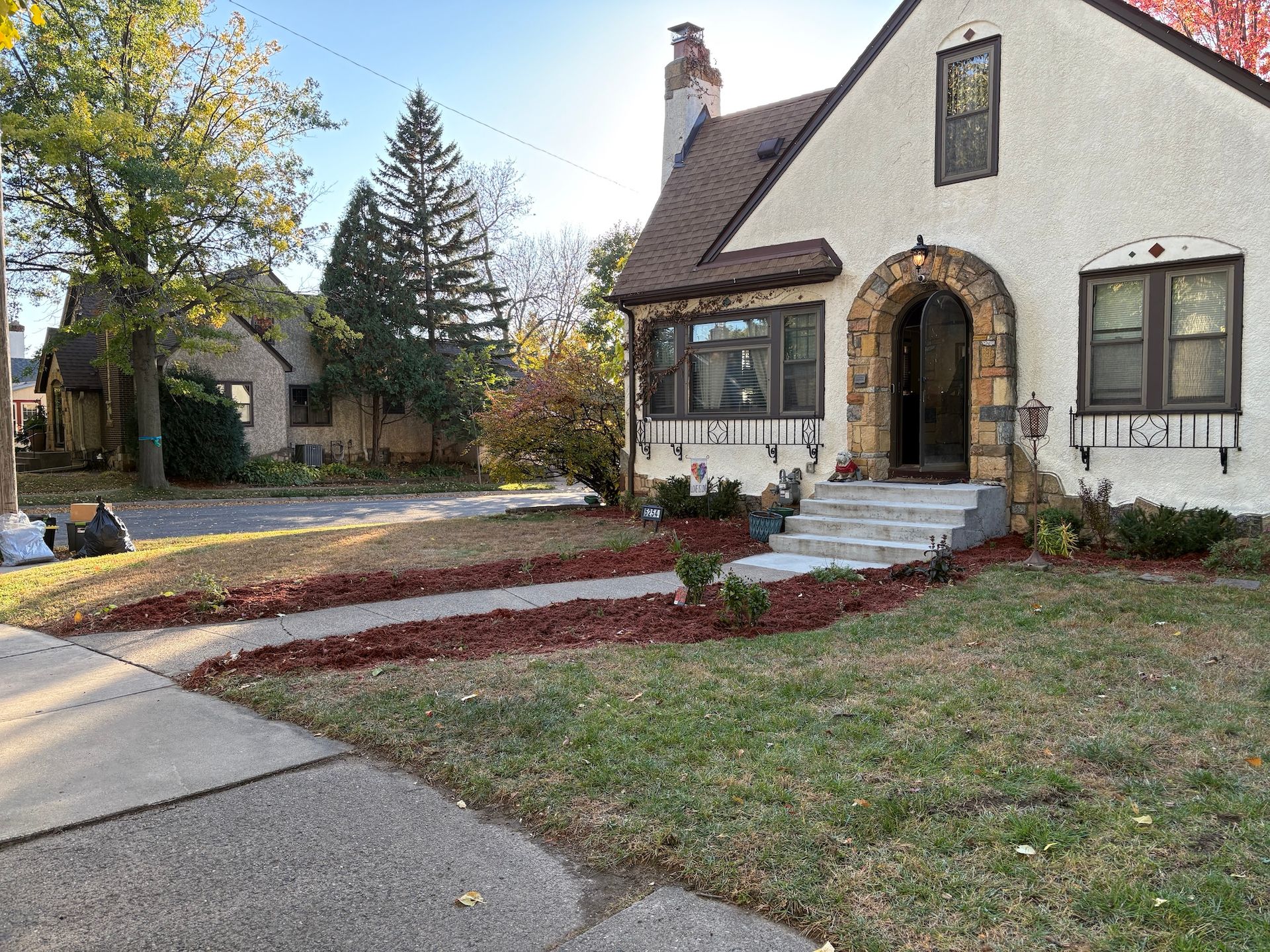 A cream-colored Tudor-style house with stone archway entry, dark shutters, and a newly mulched front walkway.