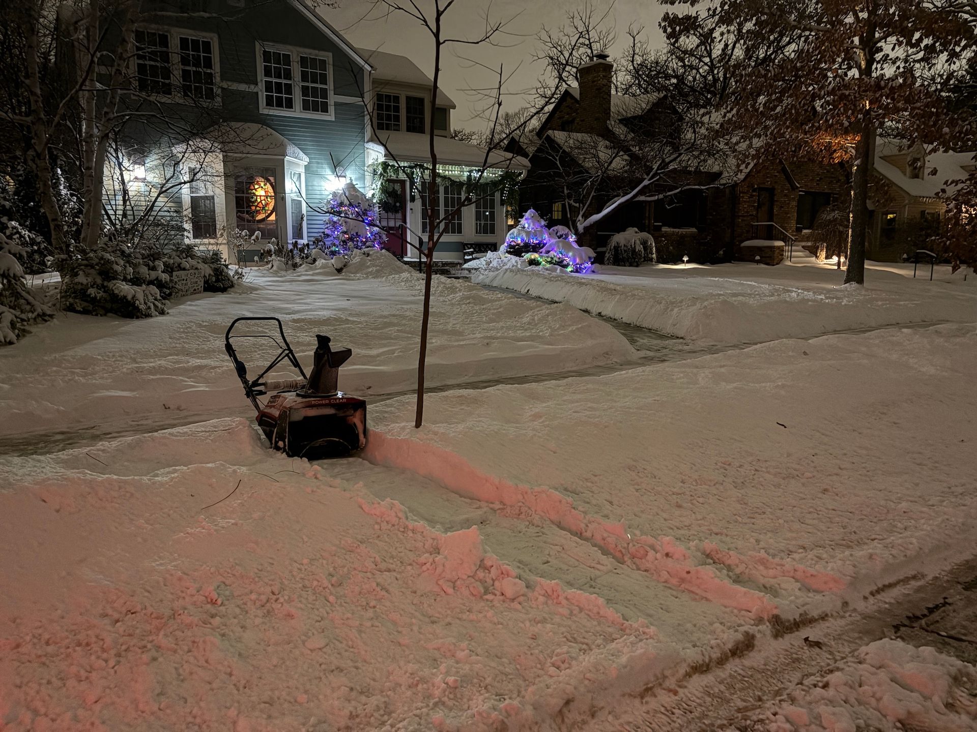A snowblower sits on a sidewalk at night, clearing a path through fresh snow in front of illuminated houses.