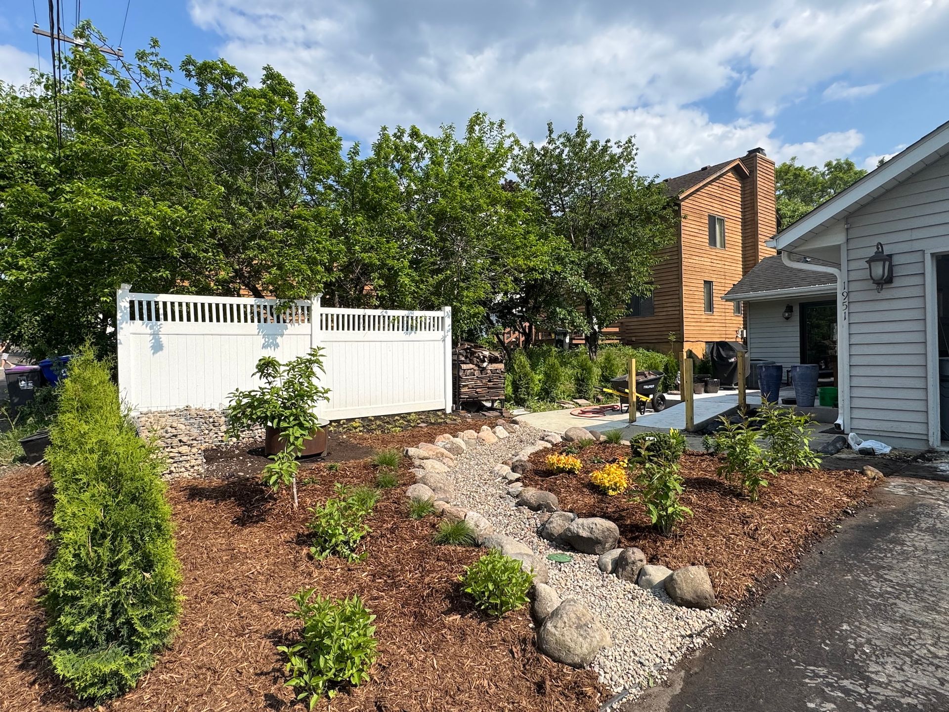 A landscaped yard with a dry creek bed, mulch, young shrubs, a white privacy fence, and a nearby house on a sunny day.