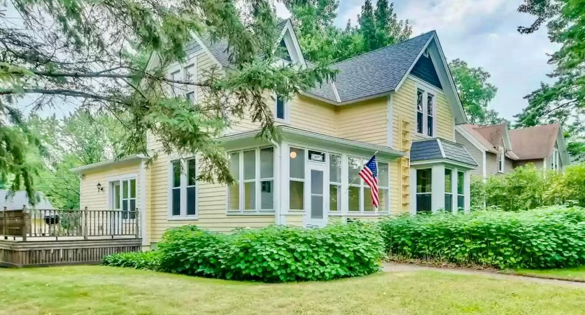 A two-story, yellow house with white trim, a wrap-around porch, and an American flag near the front entrance.