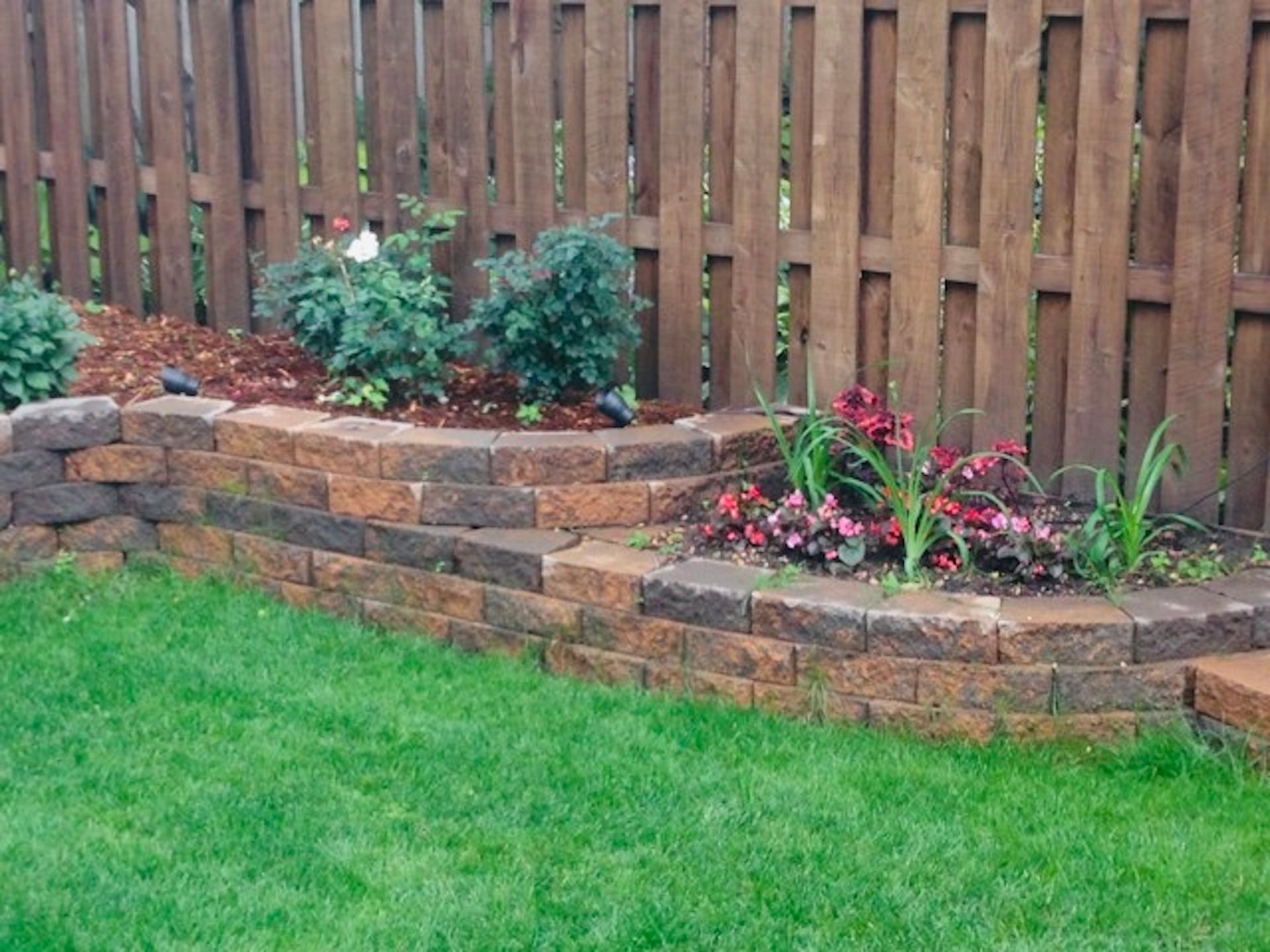 A retaining wall built from reddish-brown bricks sits in front of a wood fence, filled with various green plants.