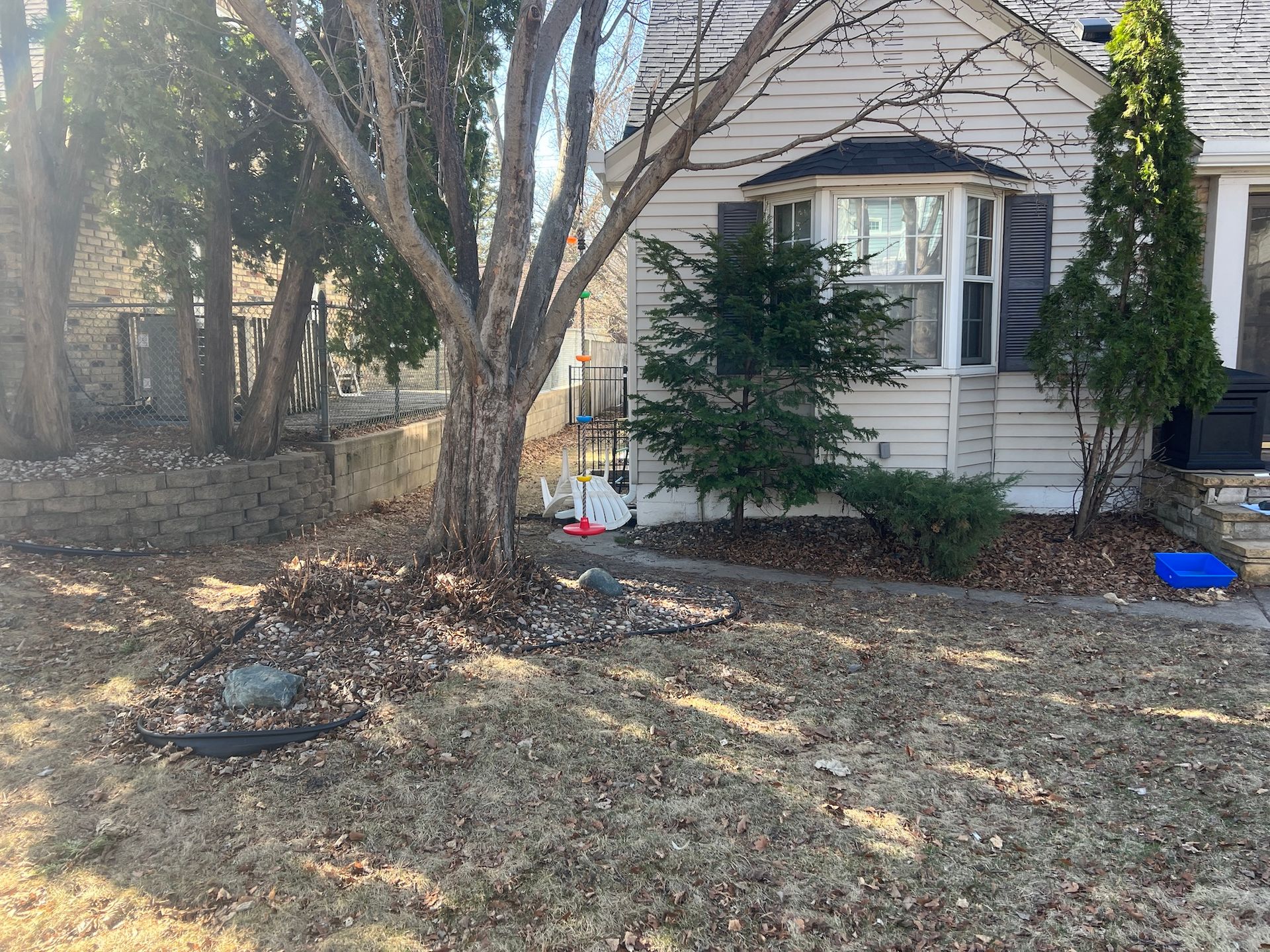 A tan house with a bay window sits behind a large tree and dry grass in a yard on a sunny day.