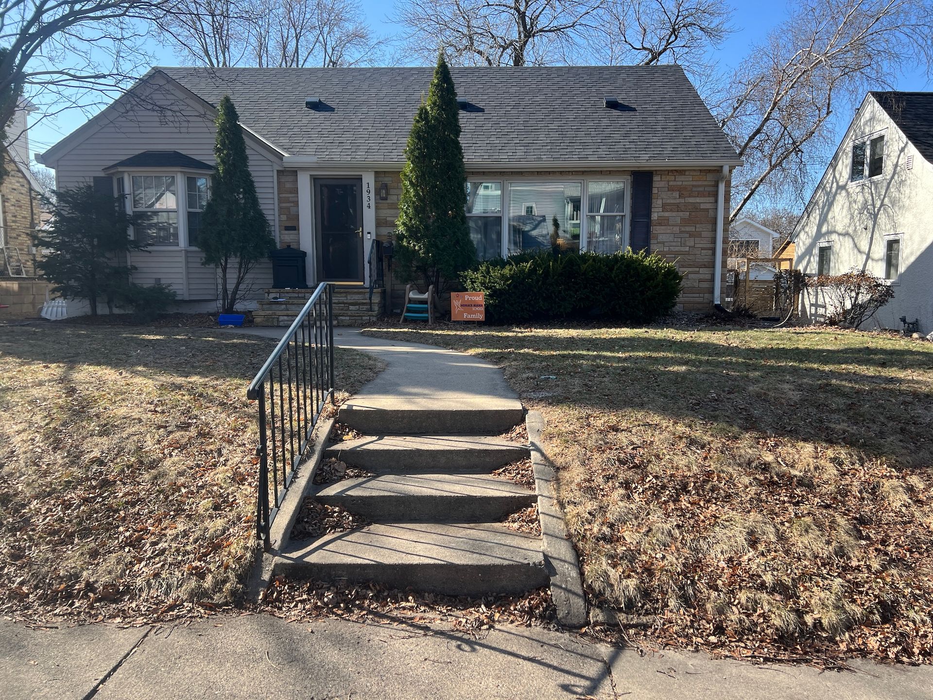 A single-story house with beige siding and stone accents, viewed from the sidewalk across a yard with concrete stairs.