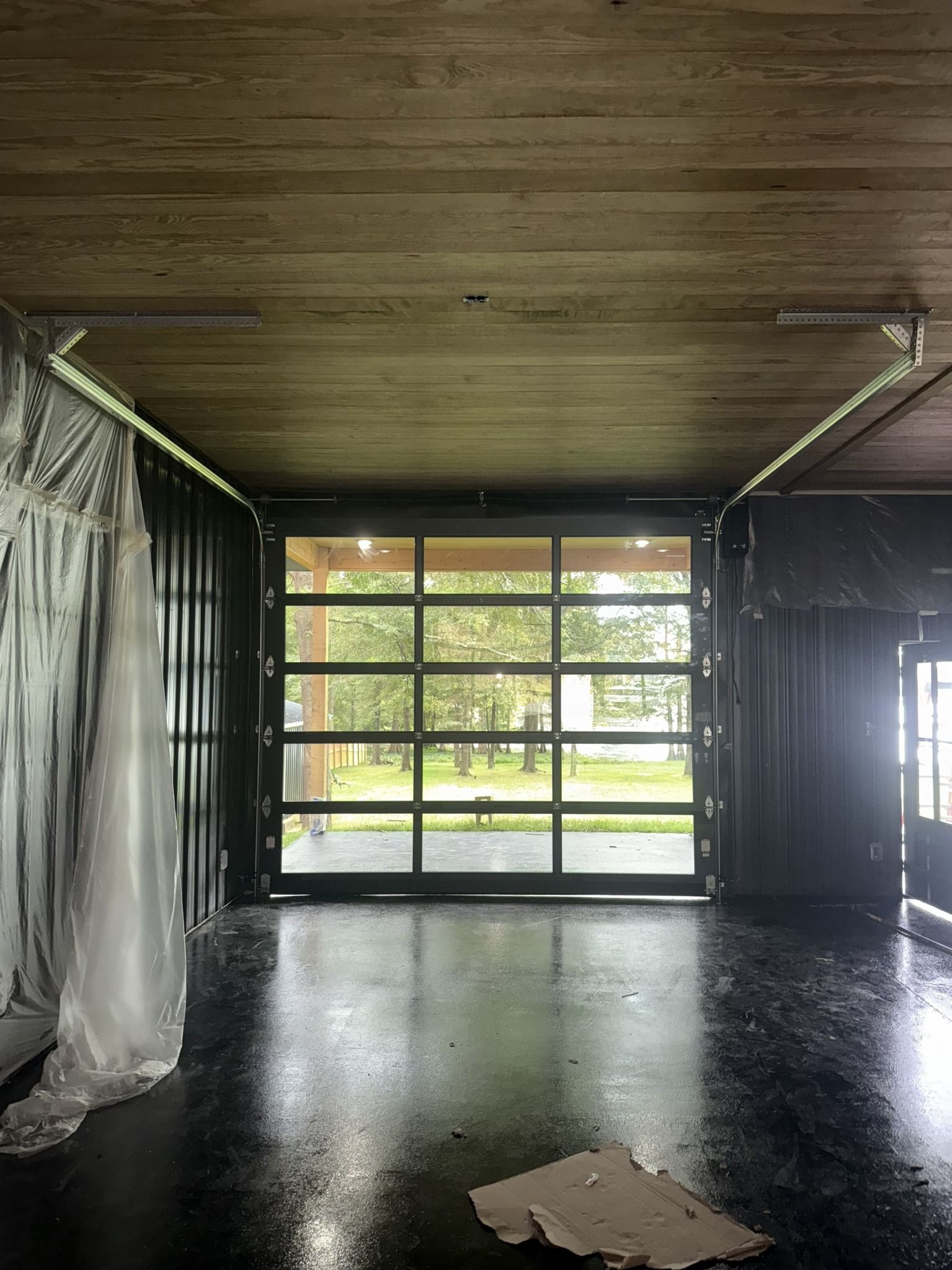 Interior of a space with a large glass door overlooking a green outdoor scene. Black floor, wooden ceiling, and dark walls.