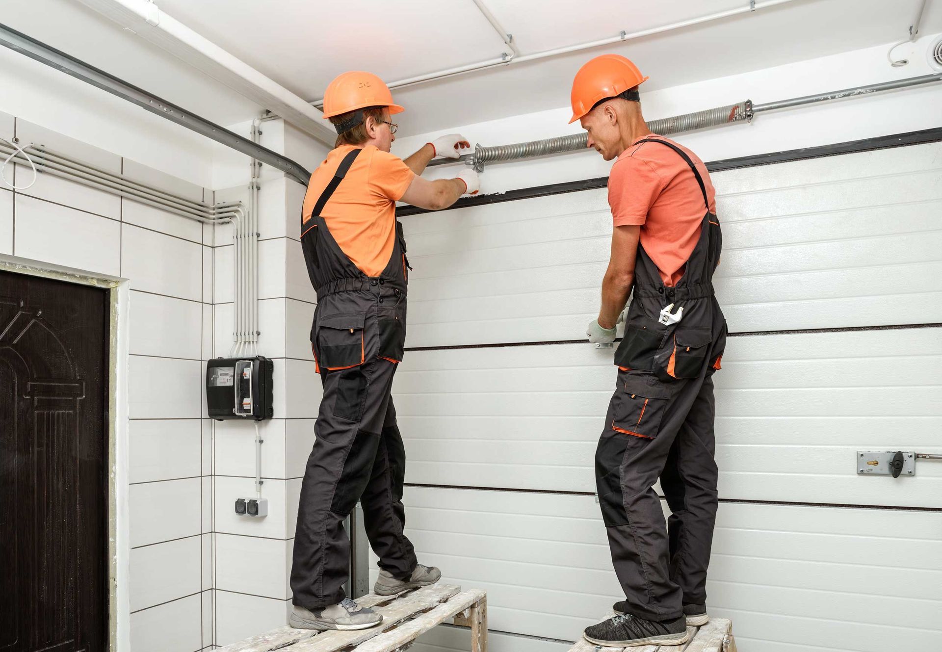 Two workers in hard hats install a metal pipe above a garage-style door indoors. Two workers in hard hats install a metal pipe above a garage-style door indoors.