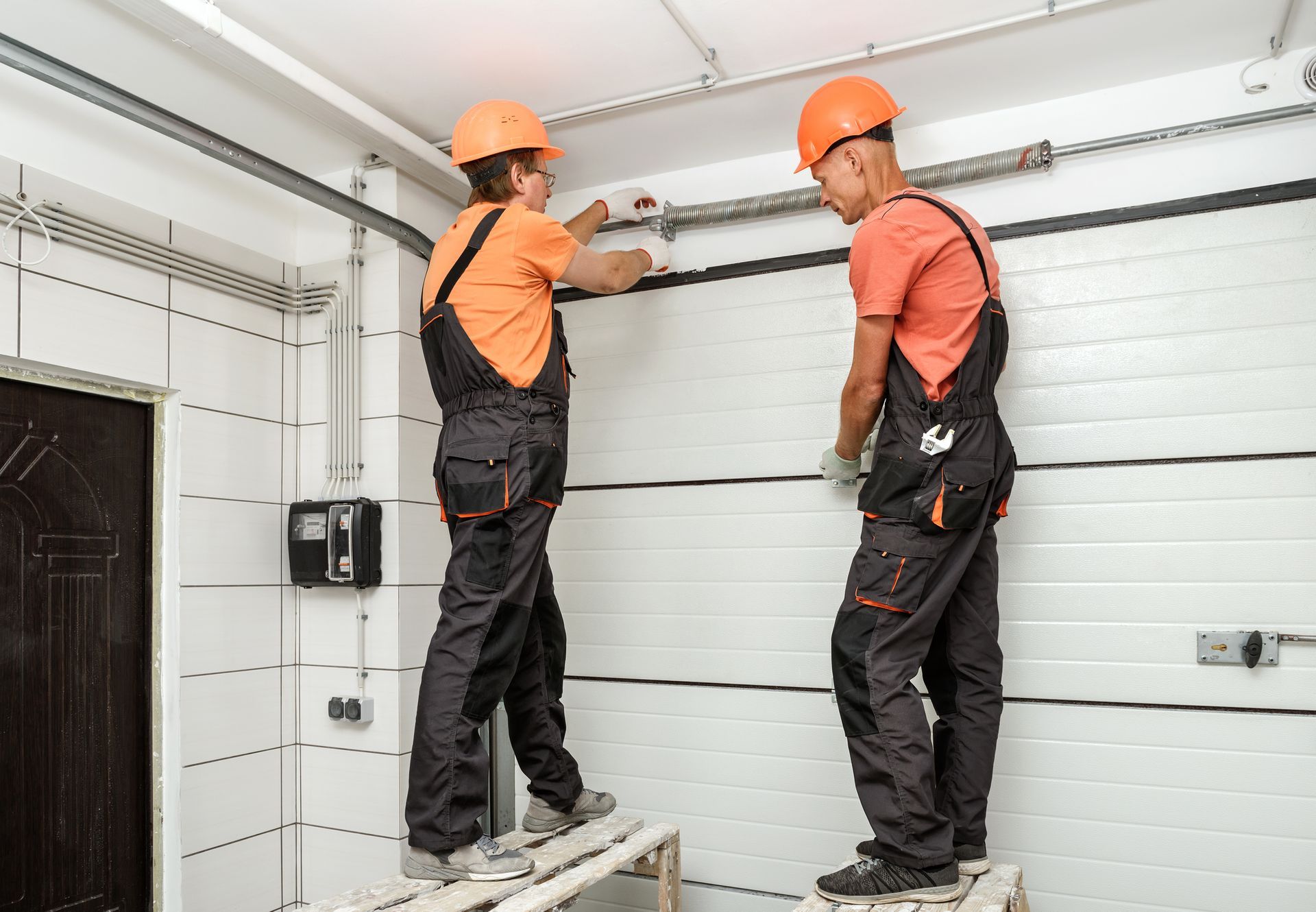 Two workers in orange shirts and helmets fixing a garage door.