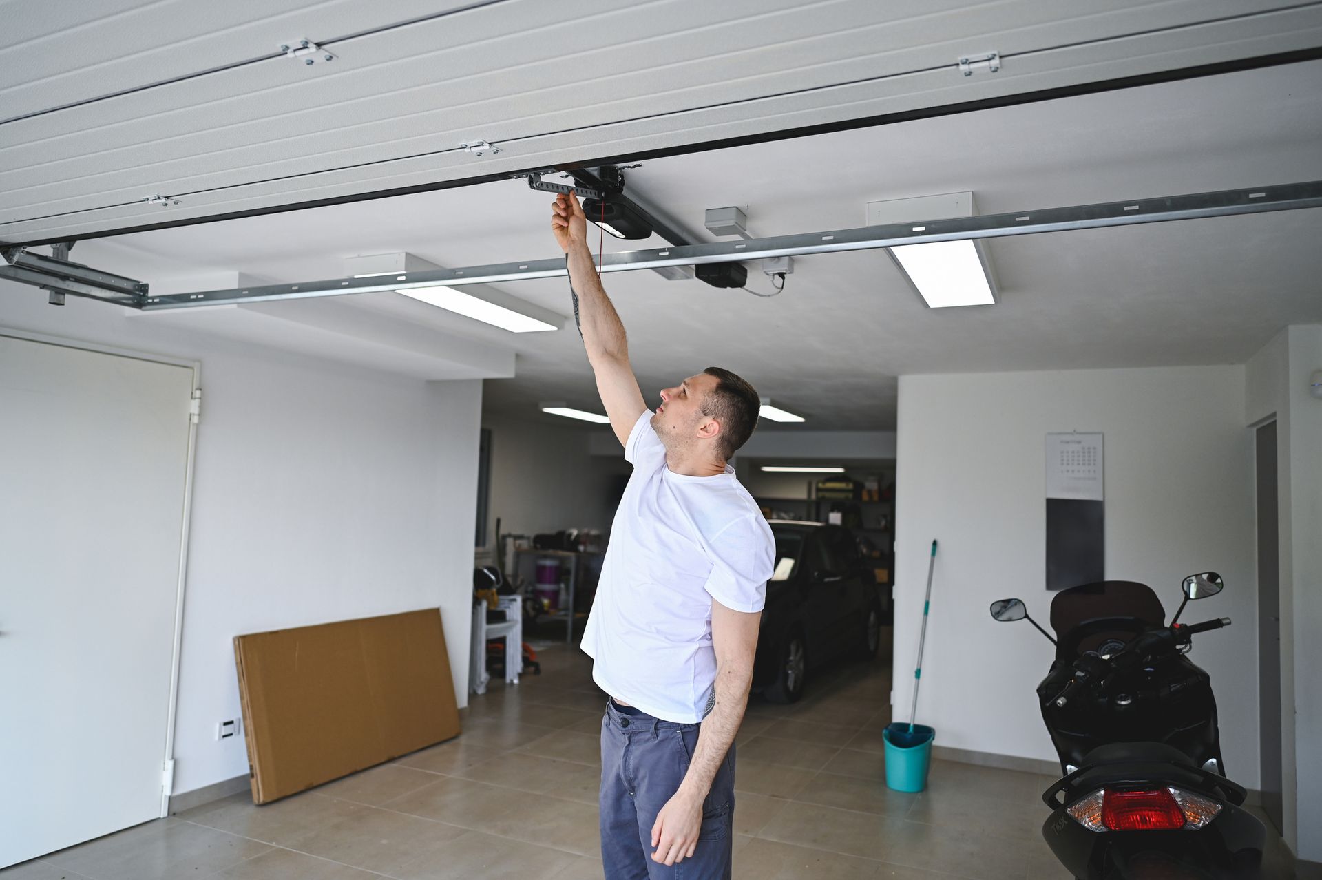 Man reaching up to a garage door opener in a garage; he is wearing a white shirt and blue jeans.