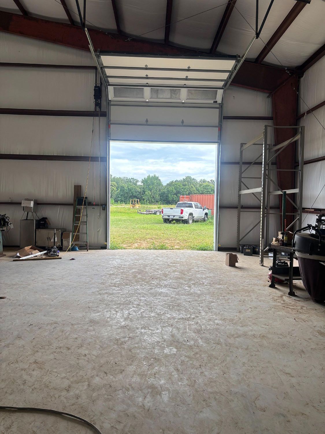 Open garage door revealing a truck in a field of grass. Steel shelving and equipment inside the building.