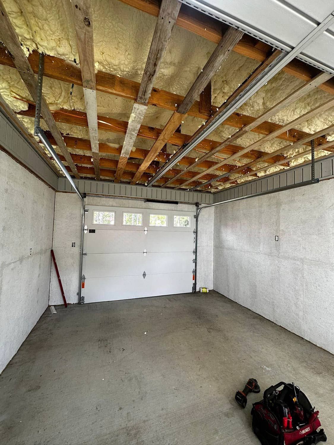 Interior of a garage with a concrete floor, white garage door, and exposed insulation on the ceiling.