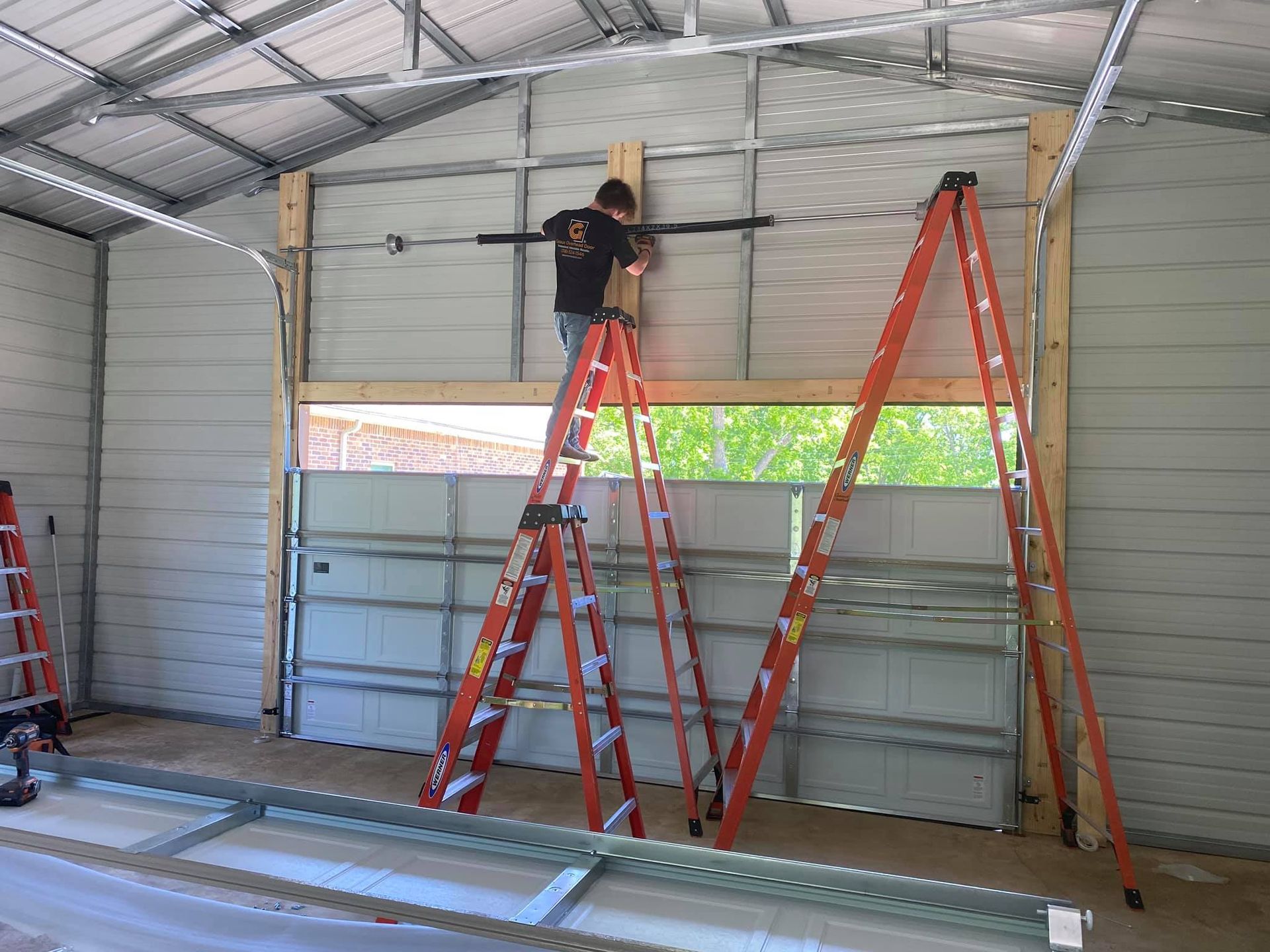 Man on ladders installing garage door opener in a metal-walled garage.