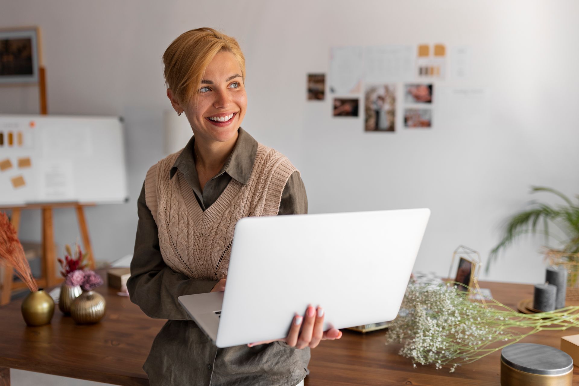 Smiling woman holding a laptop in a bright workspace with shelves, plants, and wall photos