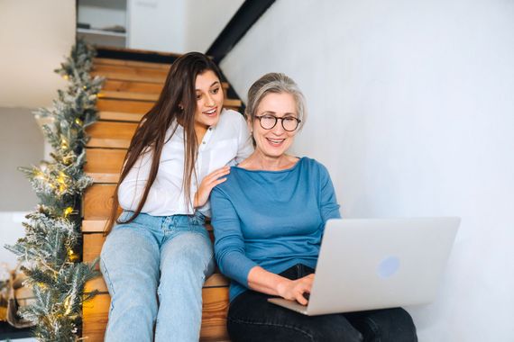 Two people sit by a staircase, smiling at a laptop in a cozy home setting.