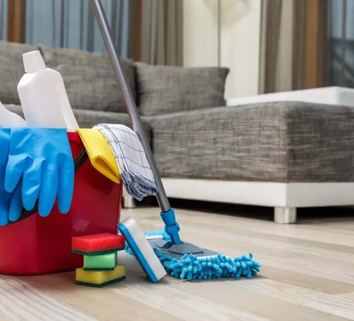 A red bucket filled with cleaning supplies, including gloves, bottles, and sponges, sits next to a mop on a wooden floor.