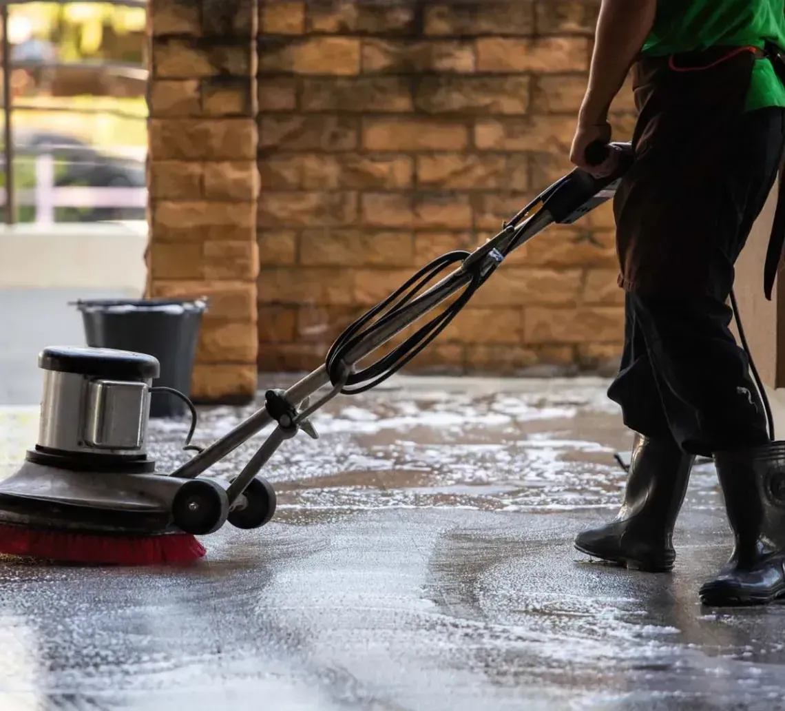 A person in a green shirt and dark apron uses a floor buffer to scrub a wet, soapy floor.