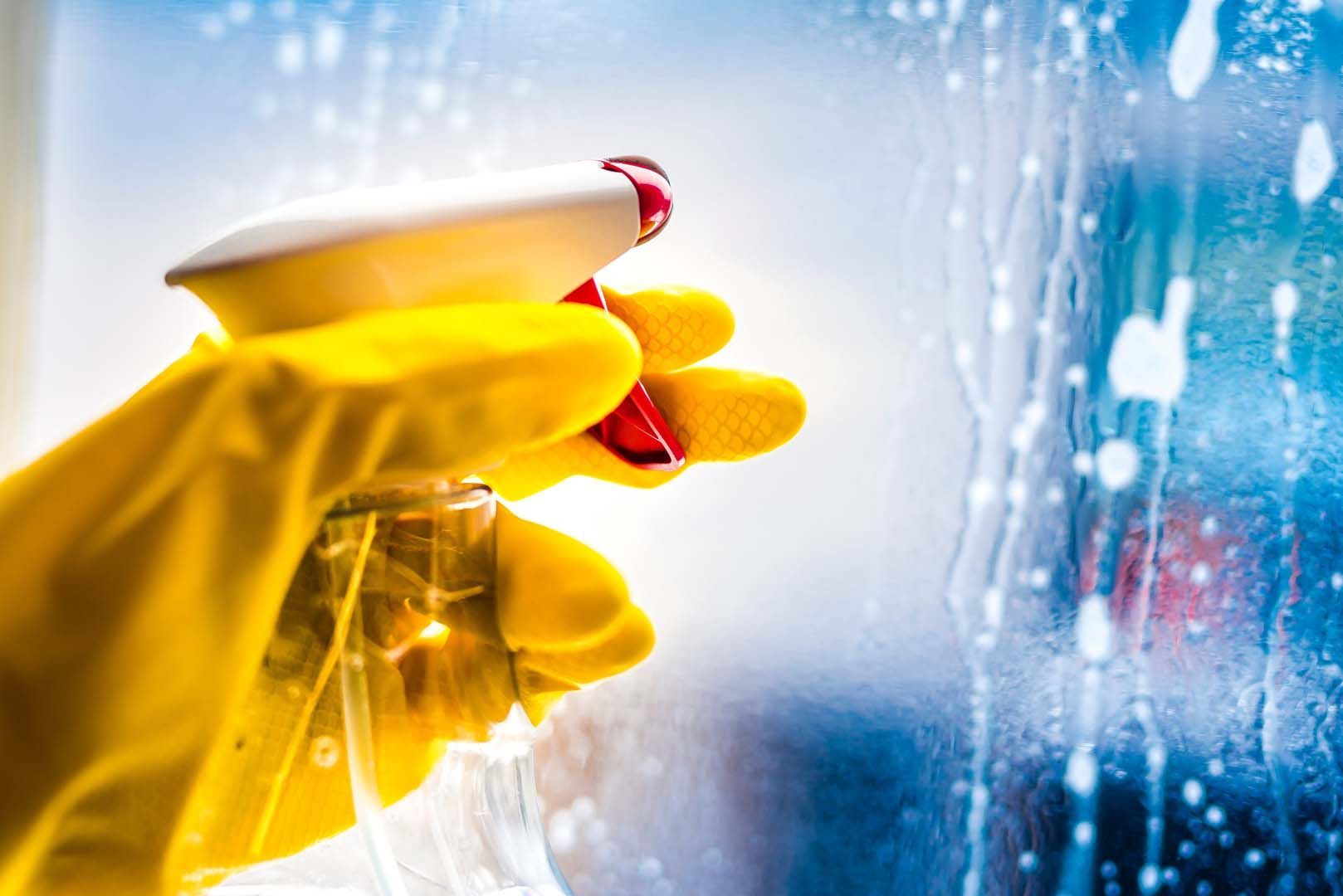 A yellow-gloved hand holding a spray bottle, cleaning a glass surface covered in cleaning solution droplets.