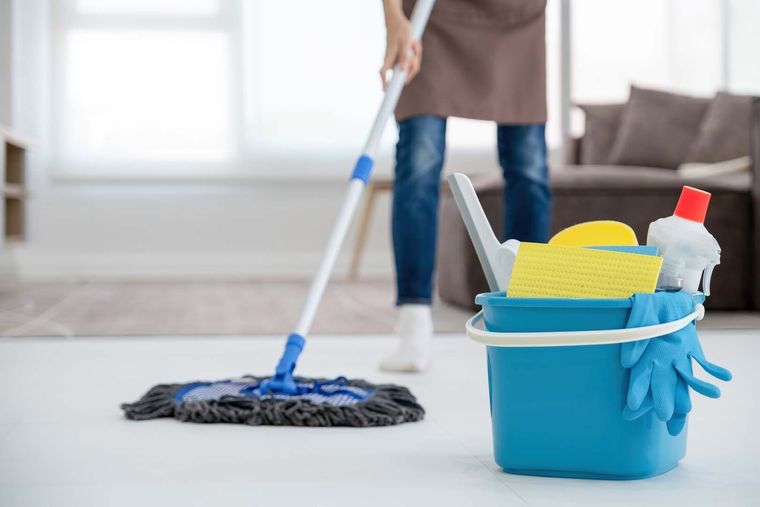 A blue bucket filled with cleaning supplies sits on a floor while a person mops the background of a living space.
