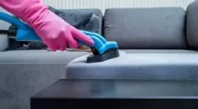 A hand in a pink rubber glove uses a blue upholstery cleaning tool to vacuum a grey sofa.