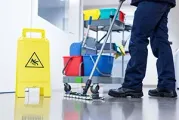 A worker in dark blue uniform mops a shiny floor next to a yellow caution sign and a rolling cleaning cart with buckets.