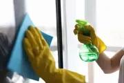 Hands in yellow rubber gloves cleaning a window with a blue cloth and a green spray bottle.