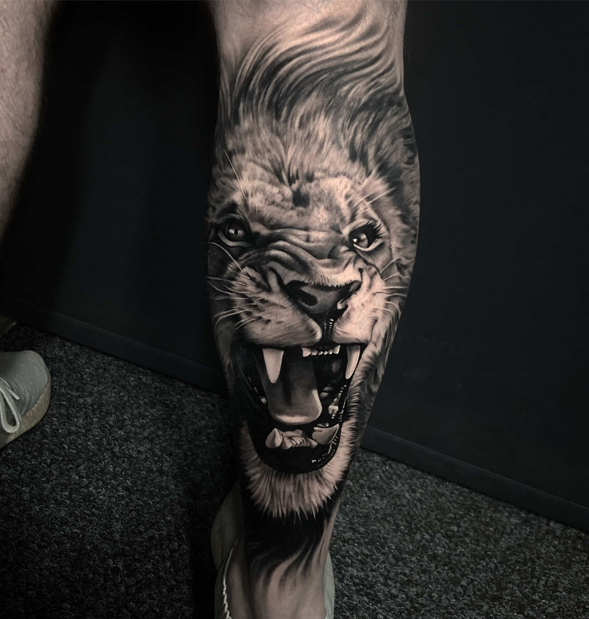 Detailed black and white tattoo of a roaring lion's face on a leg.