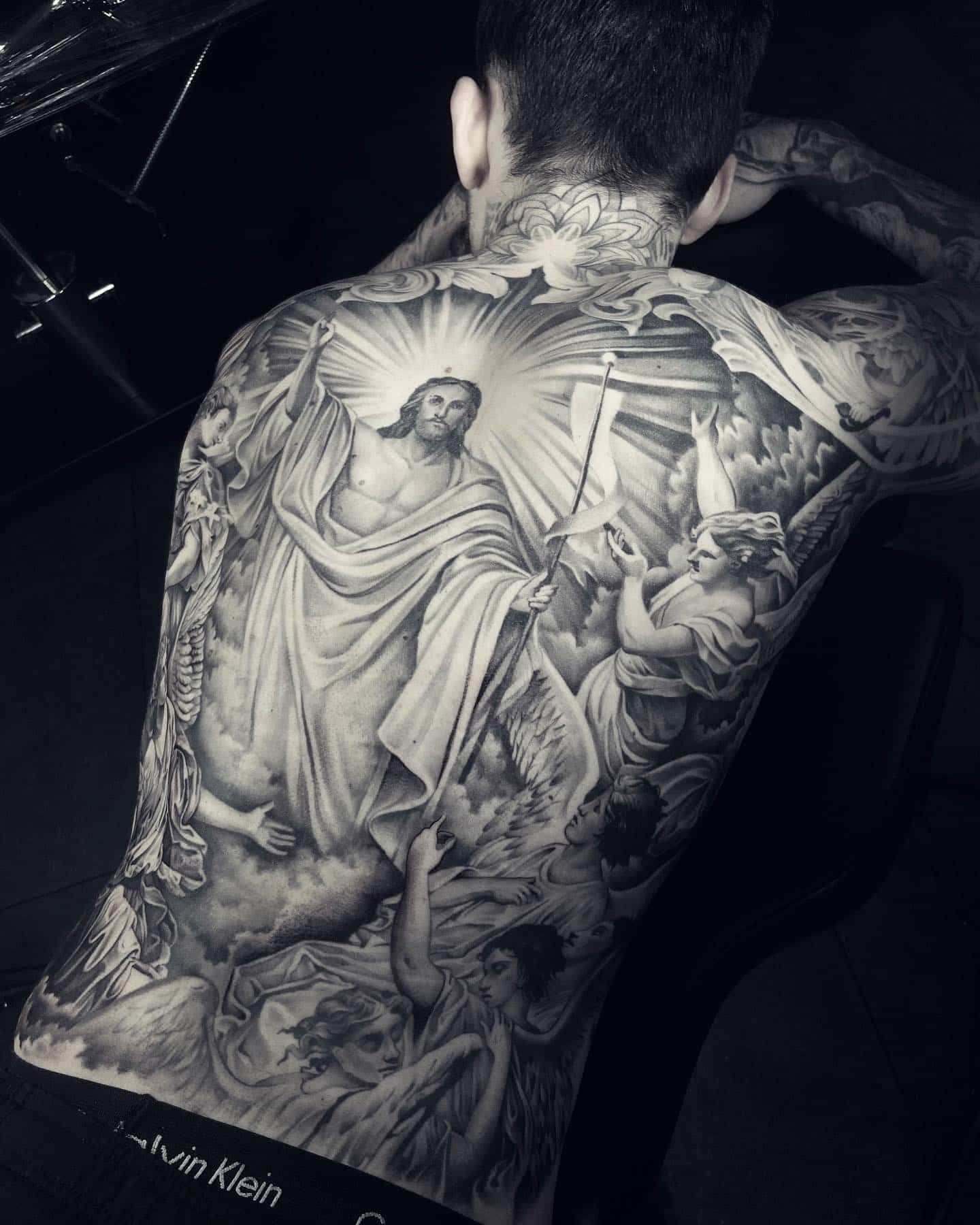 Tattoo of Jesus surrounded by angels, depicted on a person’s back; black and white.
