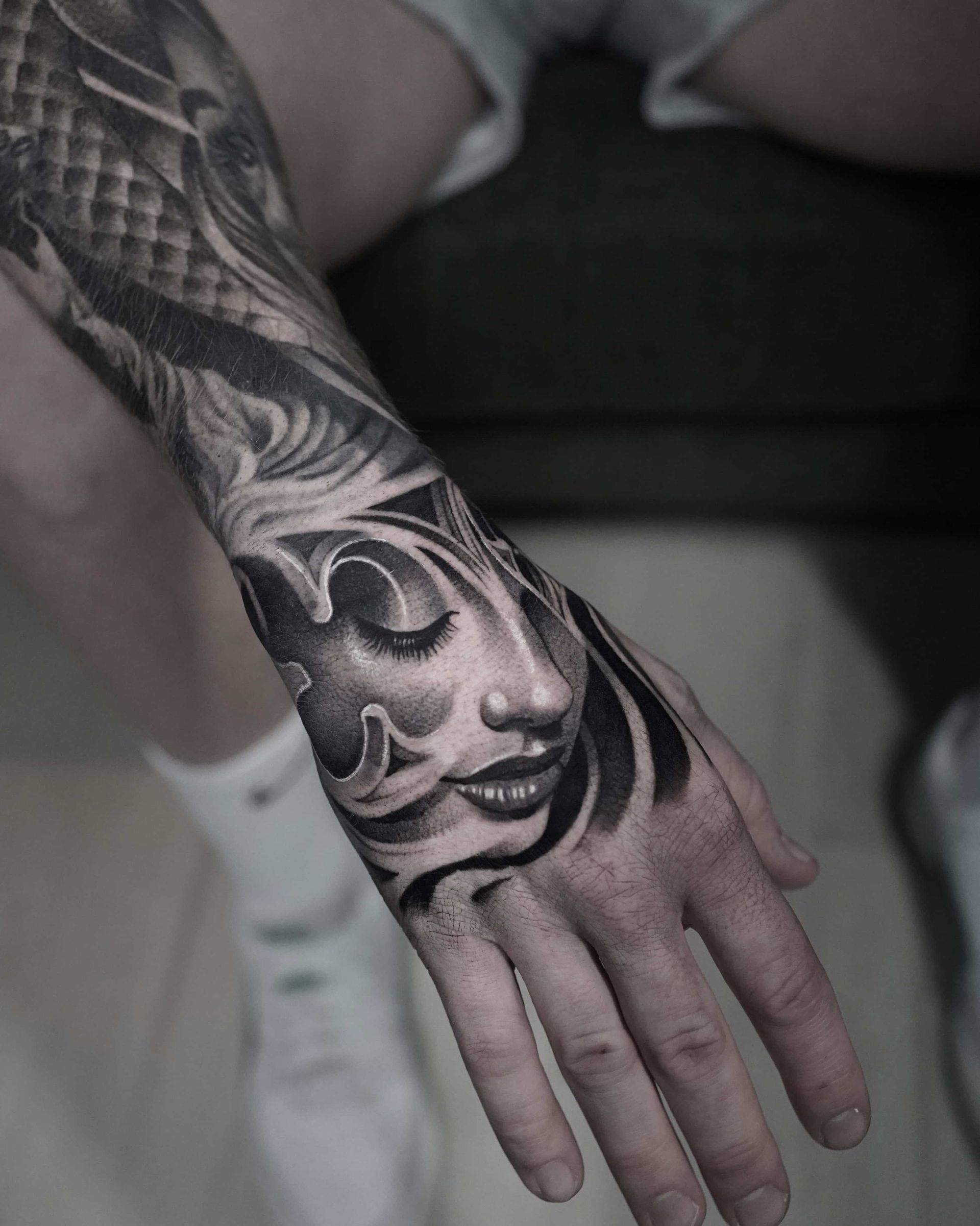 Black and gray tattoo of a serene face on a hand, arm shows ornate patterns.