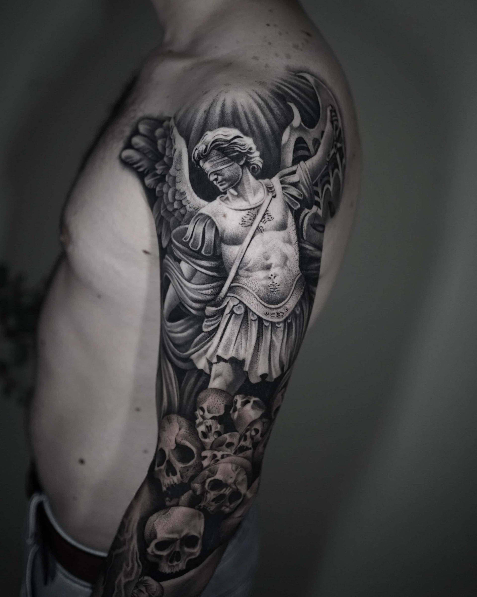 Black and white tattoo of an angel wielding a weapon, standing above skulls on a man’s arm.