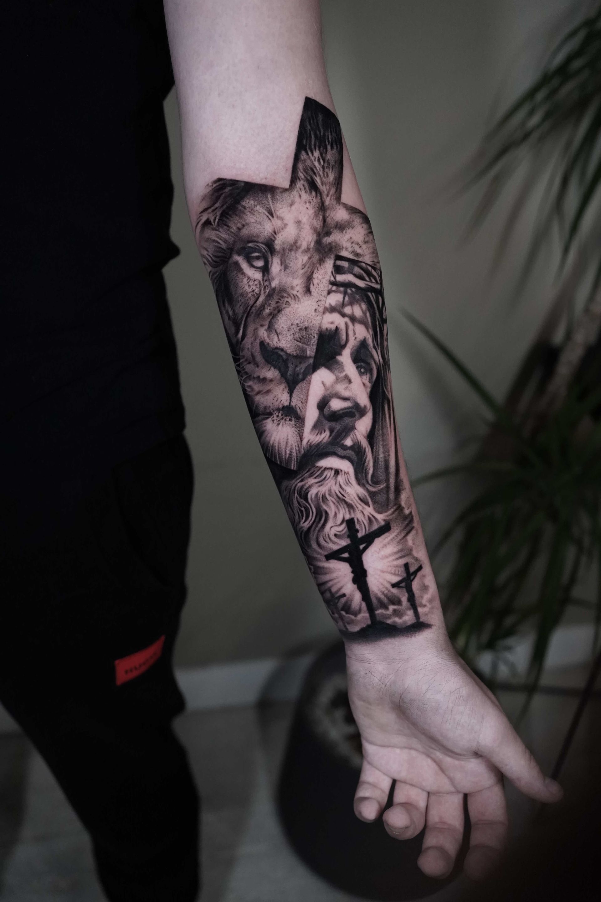 Tattoo on arm featuring a lion's head, the face of Jesus, and a cross on a hill against a sky.