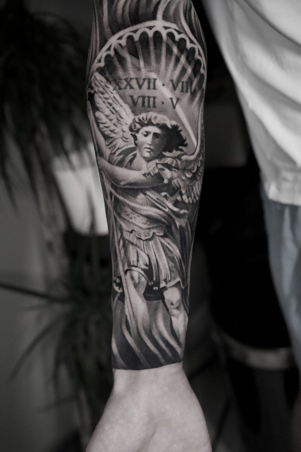 Tattoo of a black and white angel sculpture on an arm, with Roman numerals above.