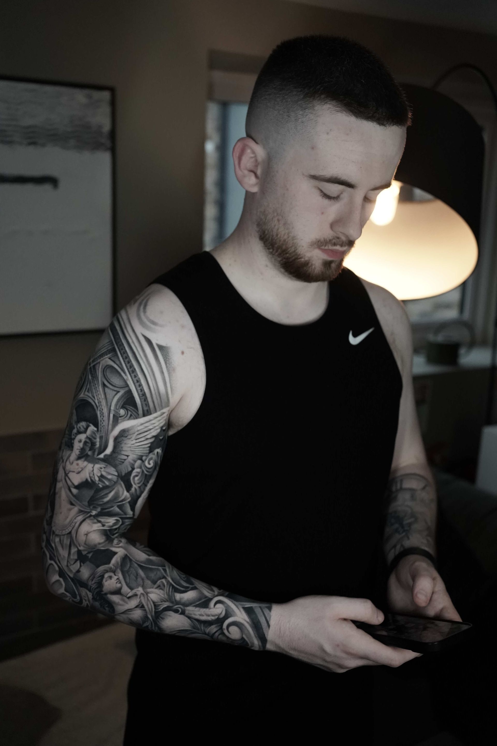 Man with arm sleeve tattoo looking down at a device; wearing a black tank top, indoors.