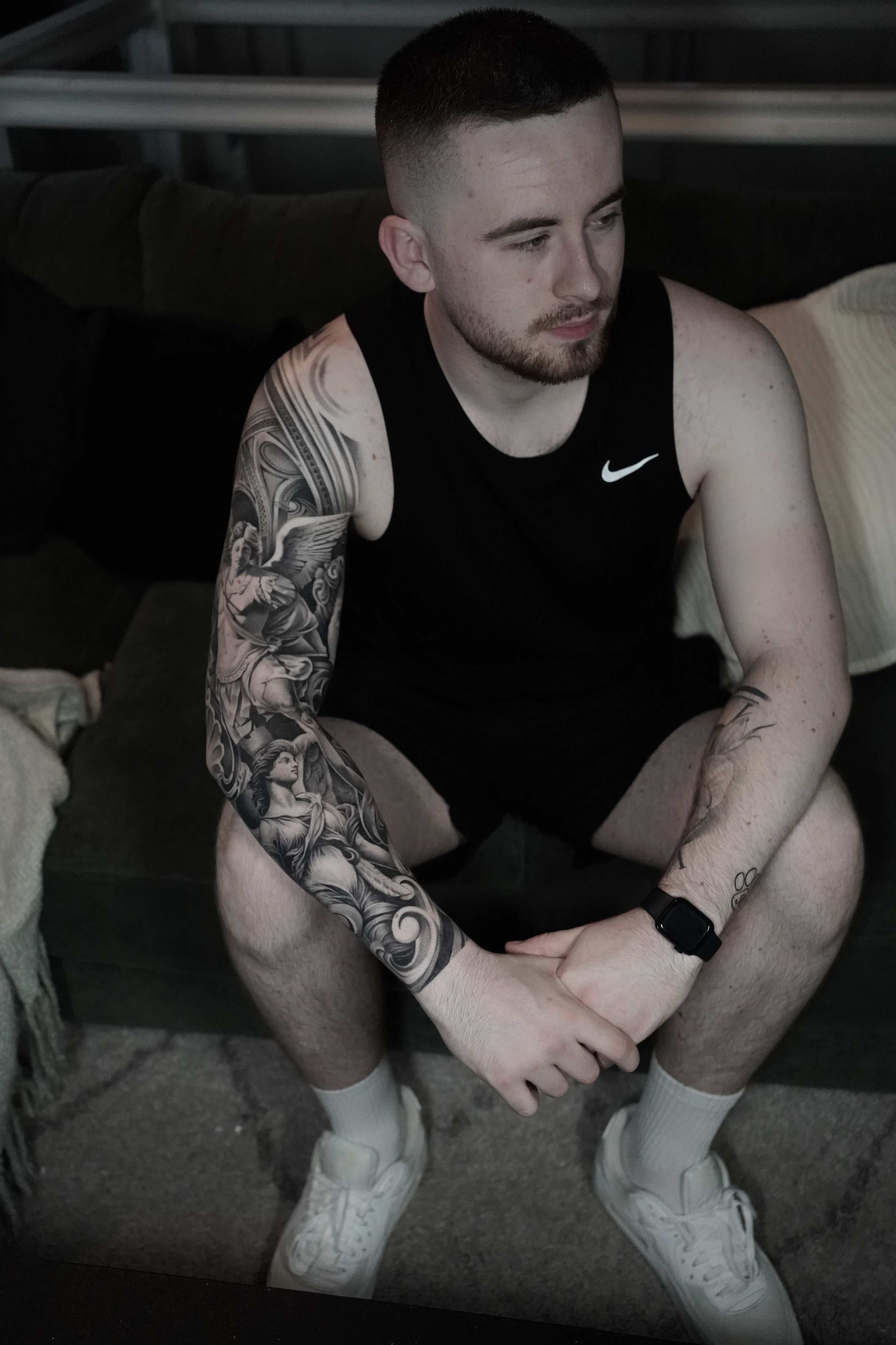 Man with arm tattoo, wearing a black tank top, sitting on a couch, looking to the side.