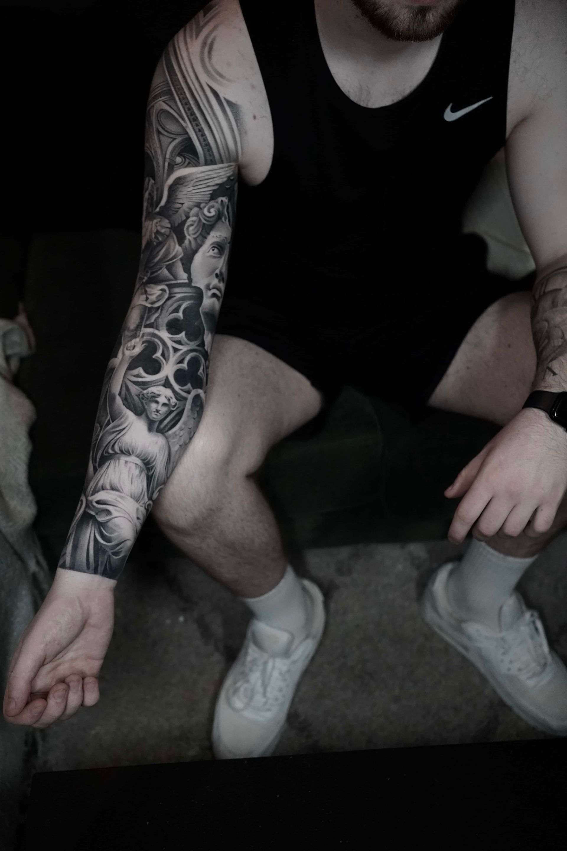 Man with a detailed black and grey arm sleeve tattoo, wearing a black tank top and white sneakers, seated.