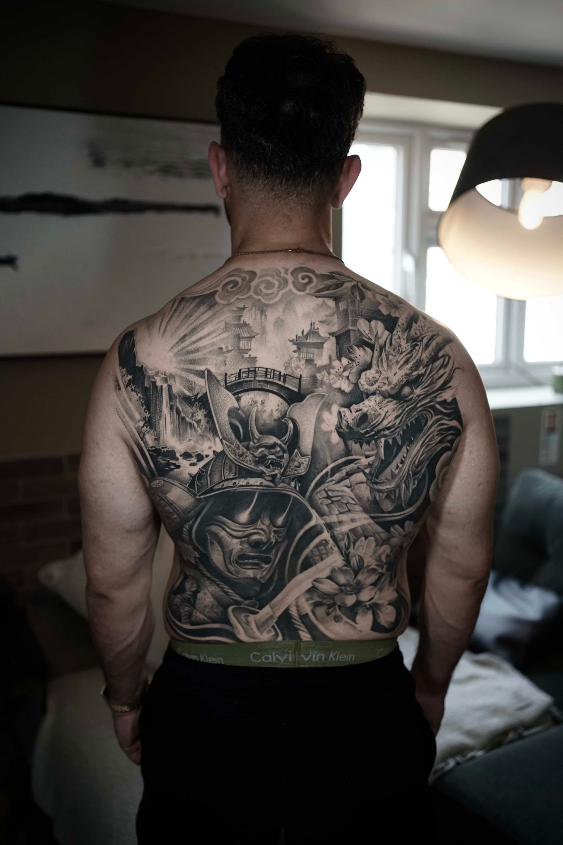 Man's back covered in intricate black and white tattoo of warriors, landscapes, and clouds.