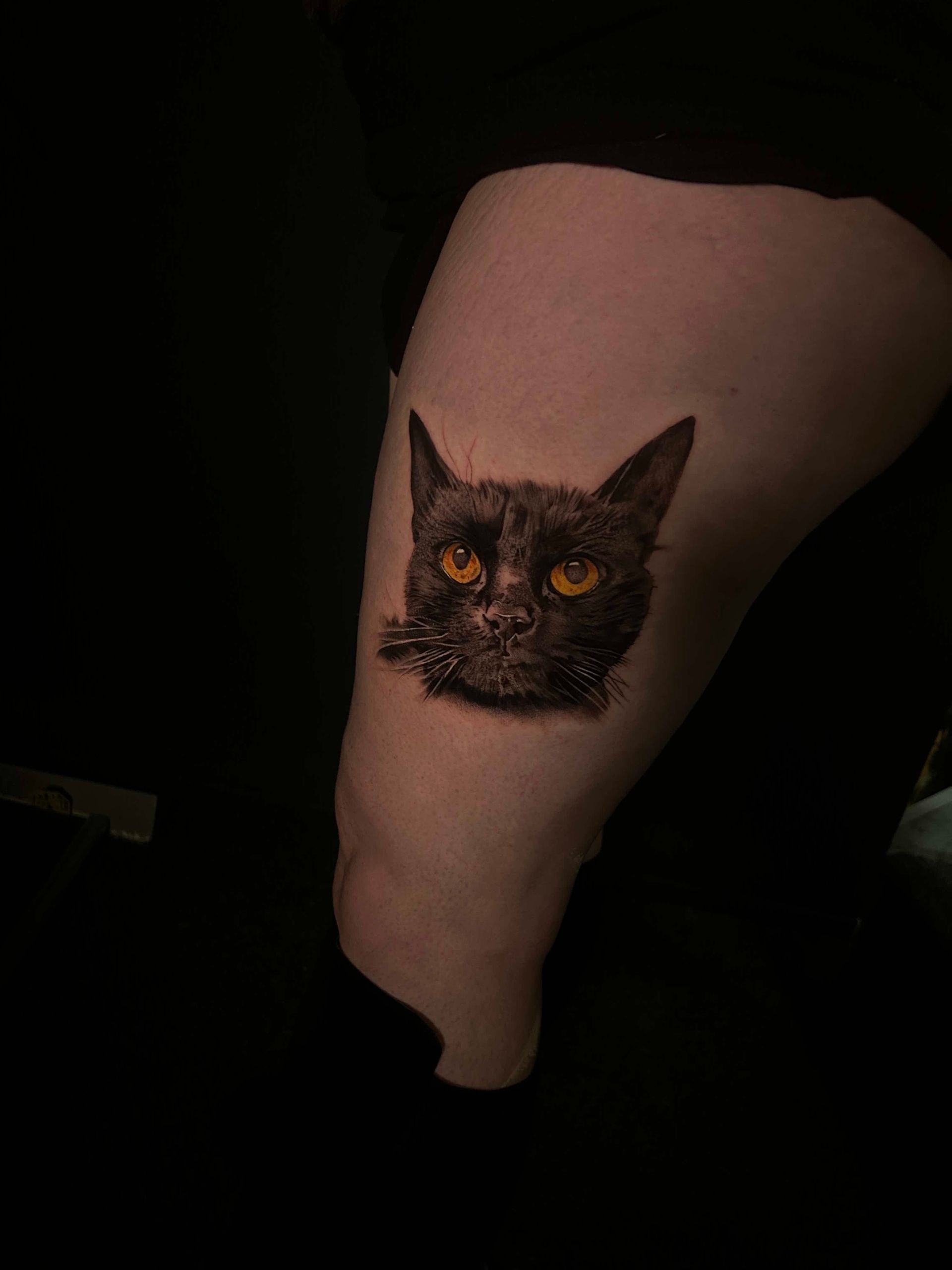 Black cat face tattoo on a thigh; orange eyes; dark fur.