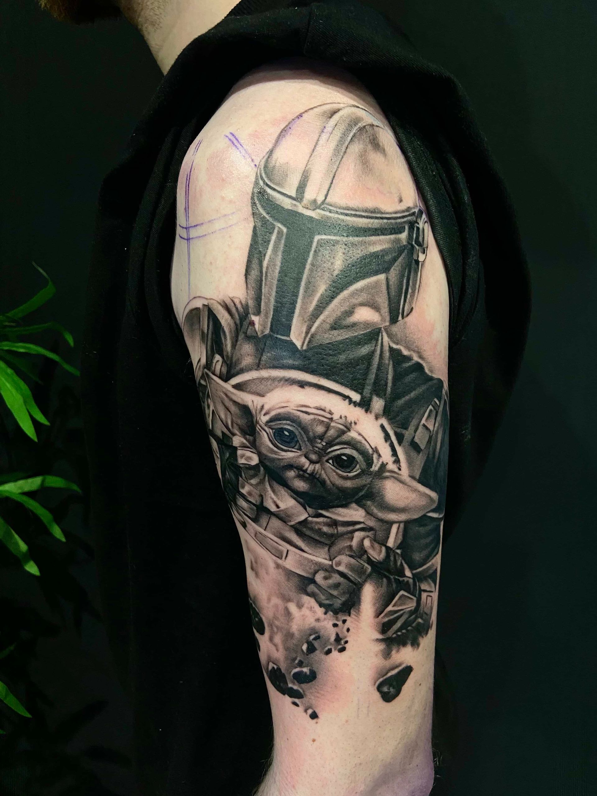 Tattoo of the Mandalorian and Grogu (Baby Yoda) on a person's upper arm, in black and gray ink.