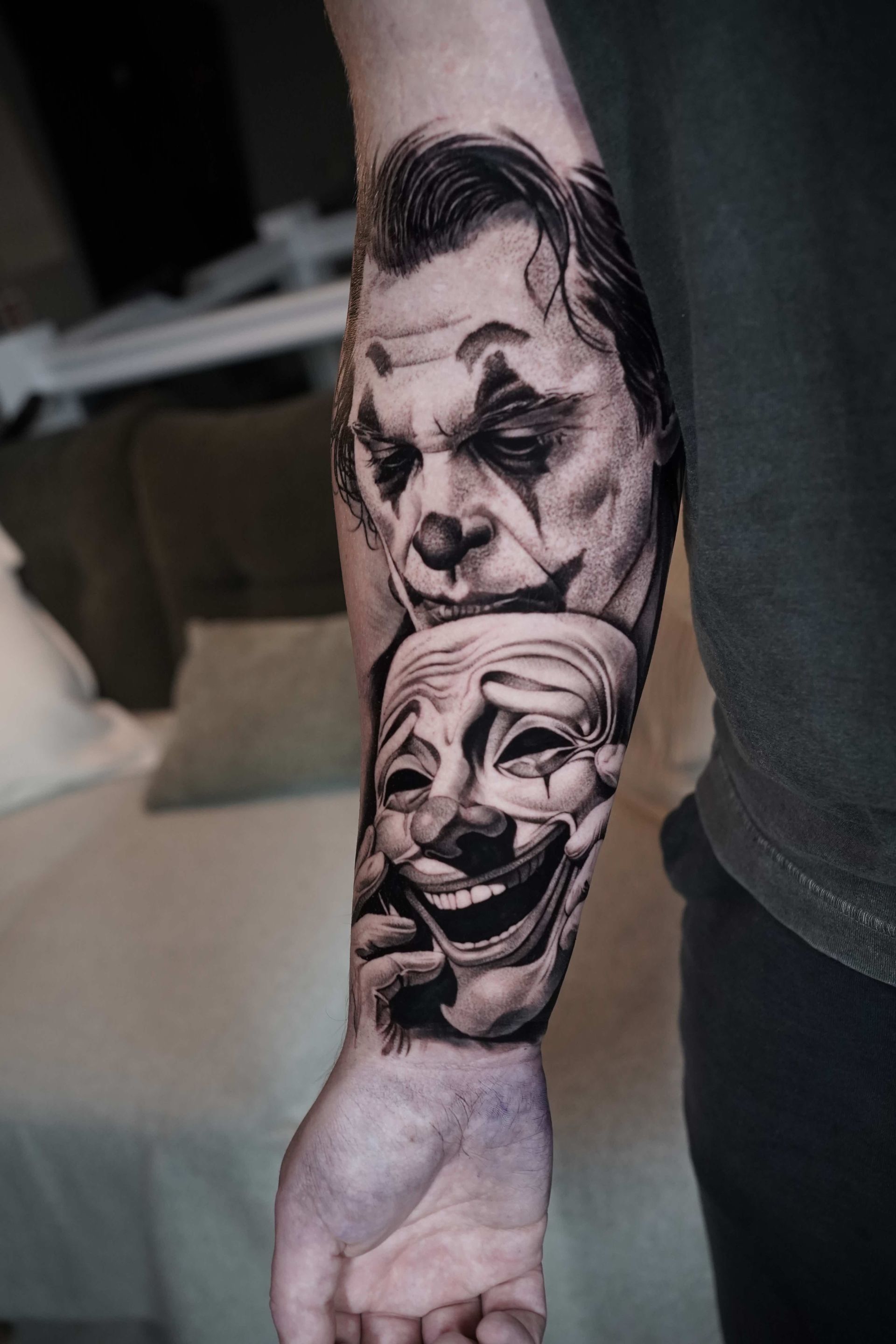 A black and white tattoo on an arm, featuring the Joker and a smiling theater mask.